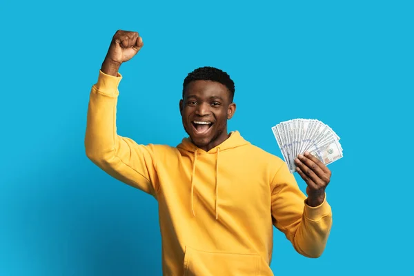 エモーショナル スリル満点のミレニアル アフリカ系アメリカ人男性 イエロー フーディーに現金ドル紙幣を振る舞い興奮させ 手を挙げ オンラインカジノで勝利を祝い 青い背景 — ストック写真