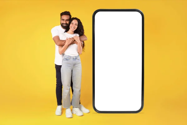 快乐的Arab夫妇拥抱着站在大智能手机旁边 在黄色工作室背景上的空白白屏上 模仿手机应用程序或提供的通信广告 全长射击 — 图库照片