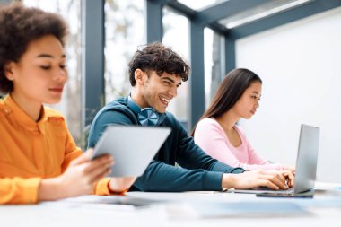 Modern oditoryumdaki masada oturan uluslararası öğrenciler, laptopta yazan ve gülümseyen adama odaklanıyorlar, sınava ya da sınıf arkadaşlarıyla kapalı alandaki derslere hazırlanıyorlar.