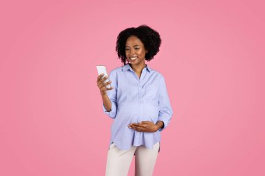 Neşeli, genç ve hamile bir kadın, sıradan, akıllı telefon kullanıyor, sosyal ağlarda sohbet ediyor, pembe stüdyo geçmişinde izole edilmiş. Uygulama, bebek beklentisi için aygıt, reklam ve teklif