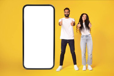 Bu başvuru formunu öneriyoruz. Heyecanlı Arap çift, büyük cep telefonunun yanında beyaz boş cihaz ekranı olan, sarı stüdyo arka planında duran başparmaklarını gösteren, tam boy, model.