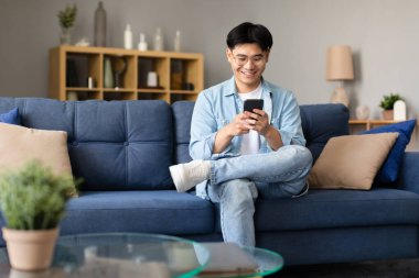Gözlüklü mutlu Asyalı adam elinde akıllı telefonuyla oturuyor, İnternet 'te geziniyor ve sosyal ağları cep telefonuyla tarıyor, uygulamaları ve mesajları evde kullanıyor. Gadgets boş zaman kavramı
