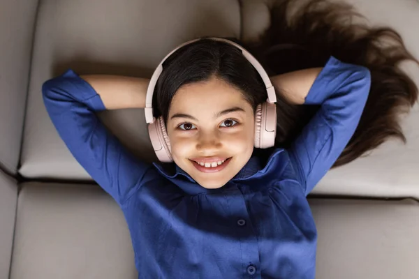 Enjoying Online Music. Cheerful kid girl listening to music wearing wireless headphones lying on couch, relaxing at home, view from above, smiling to camera. Musical fun concept