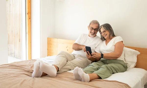 快乐的老高加索人夫妻躺在床上 在电话里聊天 在卧室里享受着早晨的愉快 周末与应用程序 小玩艺儿 关系和爱情 生活方式 — 图库照片