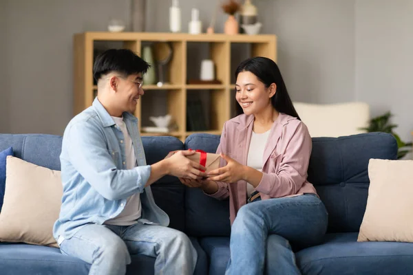 Romantic Present. Asian Young Couple Celebrating Holiday, Holding Wrapped Present Box, Husband Congratulating Wife With Gift On Birthday, Anniversary Or Family Holiday Sitting On Couch At Home