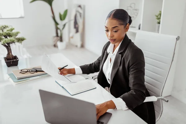 Professional recruitment concept. Black female HR manager using laptop and holding candidates CV resume, sitting in office. Businesswoman reading papers near computer at workplace