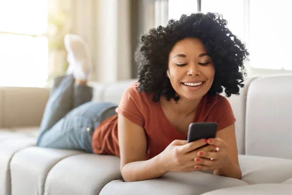 Smiling Young Black Woman Relaxing With Smartphone On Couch At Home, Happy African American Female Wearing Wireless Earbuds Using Mobile Phone, Listening Music Or Browsing Social Media, Closeup