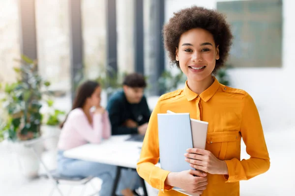 Portrait of happy black lady university student posing with notepads and smiling while her classmates studying on background in modern audience interior