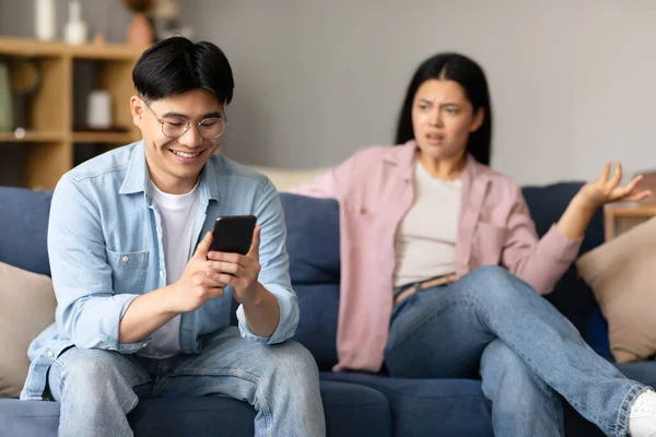 Phubbing Concept. Happy Asian Husband Texting On Smartphone Ignoring His Displeased Wife, While Woman Suspecting Infidelity Looking At Him, Sitting On Couch At Home. Selective Focus