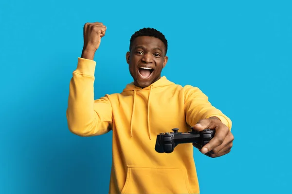 ジョイスティックコンソールジェスチャー クレンチフィスト ブルースタジオのバックグラウンドでビデオゲームをする黄色いパーカーを着た幸せなハンサムミレニアルブラック男 テクノロジーへの参入 — ストック写真