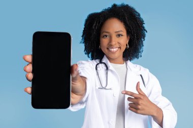 Beyaz önlüklü bayan doktor, mavi stüdyo arka planında izole edilmiş beyaz ekranlı akıllı telefona işaret ediyor. Aygıt, iş uygulaması, profesyonel tavsiye tıbbi yardım