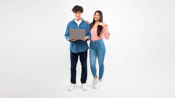 Ecommerce. Smiling Young Couple Enjoying Online Shopping On Laptop Holding Credit Card, Promoting Bank Offers and Internet Payment Services Standing Over White Studio Background. Panorama