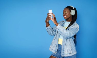 Neşeli, şık, güzel, Afro-Amerikan bayan 20 'li yaşlarda, cep telefonu ve modern kablosuz kulaklıkla müzik dinliyor, selfie çekiyor, görüntülü sohbet ediyor, mavi arka plan, fotokopi çekiyor.