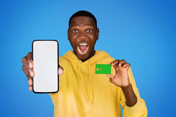 令人高兴的是 千禧年的非洲裔美国人 张开嘴 带着空白屏幕的电话 信用卡 在蓝色的工作室背景下与世隔绝 推荐应用程序 网上购物 — 图库照片