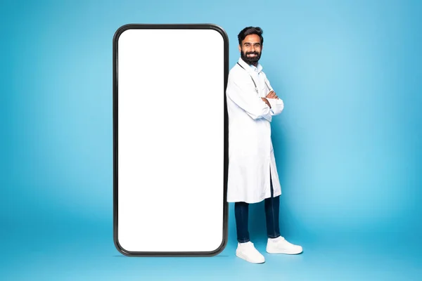 身穿白衣的印度男医生靠在装有白屏的巨大智能手机上 站在蓝色背景之上 进行模仿 宣传医疗移动应用的专业医生 — 图库照片