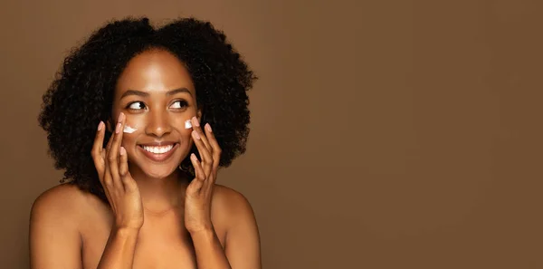 Happy pretty topless young black woman with smooth youth-looking skin apply hydrating, moisturizing organic beauty product cream on face and smiling, brown background, copy space. Skin care routine