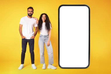 Sıradan bir ortadoğu çifti, boş ekranlı büyük akıllı telefonun yanında durup kameraya, reklam teklifine ya da mobil telefon uygulamasına bakıyor, sarı arkaplan üzerinde. Tam boy çekim