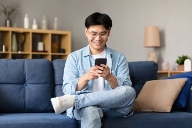 Mobil Uygulama. Mutlu Koreli genç adam akıllı telefon mesajları kullanıyor, gözlük takıyor ve evdeki koltukta oturup eğlenmek için başvuru yapıyor. Erkek Mesajlaşması Cep Telefonu Gadget İçerde İnternet Taraması