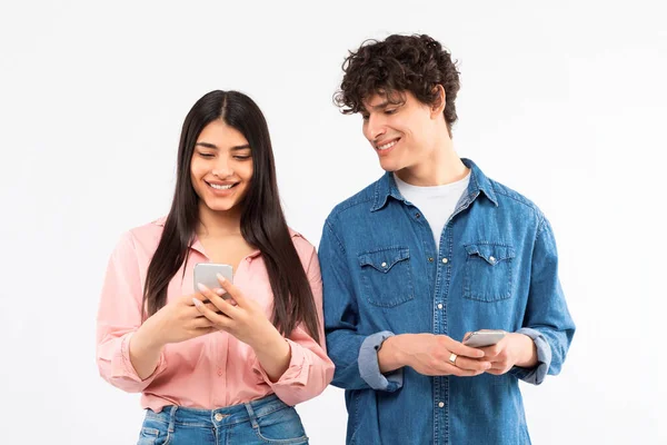 stock image Gadgets, Applications And Communication. Smiling Young Couple Using Cellphones, Guy Peeking At Girlfriends Smartphone Posing On White Background. Studio Shot. Great Mobile Offer Concept