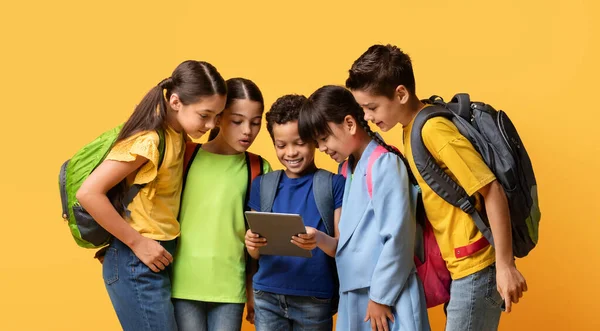 バックパックを持つ多文化の子供たちのグループは タブレットを調べます 子供のコンピュータゲーム ソーシャルネットワーク 教育アプリのコンセプト 黄色の背景にパッドを持つ少女と少年 コミュニケーション技術 — ストック写真