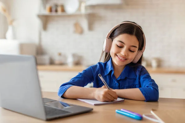 Home schooling. Cheerful middle eastern girl studying online, using laptop and headset indoor, copy space. Cute arabic preteen school kid having online lesson, drawing sitting at kitchen interior