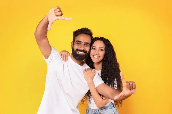 Cheerful Middle Eastern Couple Framing Themselves For Photo, Posing Together In Casual, Making Selfie Finger Frame And Having Fun Over Yellow Background, Smiling At Camera. Studio Shot