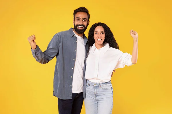 Yes, We Did It. Joyful Young Middle Eastern Couple Shaking Fists Smiling To Camera, Celebrating Their Common Victory And Success, Embracing Standing Over Yellow Background. Joy, Luck Celebration