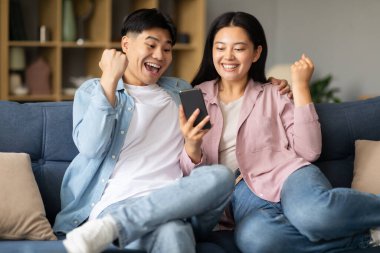 Mutlu Asyalı çift akıllı telefona bakarken başarıyı kutluyor ve evde el kol hareketi yapıyor, heyecanlı genç eşler telefonda tezahürat yapıyor, duygusal olarak iyi haberlere yumruk kaldırıyor.