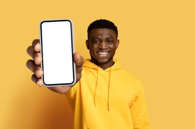 Sarı kapüşonlu neşeli yakışıklı genç adam beyaz ekranlı büyük bir telefon gösteriyor, güzel bir online teklif öneriyor, mobil uygulama, sarı arka planda izole edilmiş, kopyalama alanı, modelleme alanı,