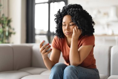 Genç Afrikalı Afro-Amerikalı kadın koltuğunda oturup akıllı telefon ekranına bakarken, Milenyum siyahi kadının evde dinlenirken önemli bir çağrı ya da SMS beklemesinden sıkıldı.