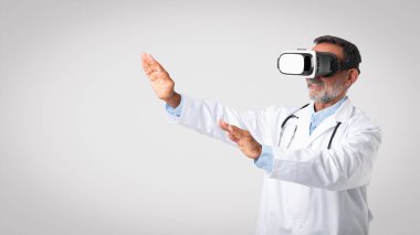 Beyaz önlüklü ve kameralı beyaz doktor terapisti gri arka planda, stüdyoda ve panoramada izole edilmiş boş alanı kontrol ediyor. Tıp, sağlık, mesleki çalışma, bilim