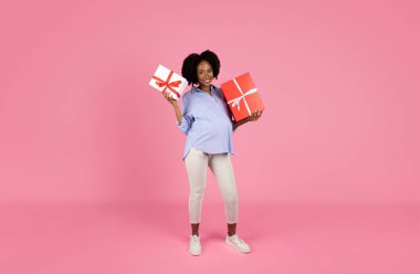 Geniş göbekli, hoş, genç, siyah hamile bir bayan, pembe stüdyo arka planında izole edilmiş, cinsiyet partisinde hediyeler sunan kutular gösteriyor. İlan et, teklif et, bekle çocuk, tatilin kutlu olsun.