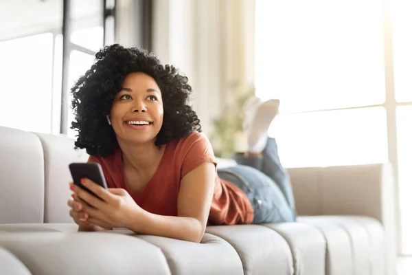 Home Relax. Portrait Of Dreamy Young Black Woman Listening Music On Smartphone, Smiling African American Lady Wearing Wireless Earbuds Lying On Couch, Enjoying Favorite Songs, Resting In Living Room