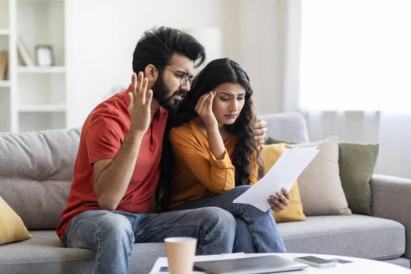 Bad News. Portrait of upset young indian couple reading documents at home, sad unhappy eastern spouses sitting on couch, checking bills, reading notification letter from bank, free space