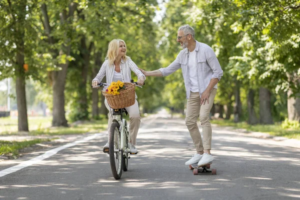 在公园的路上 活跃的情侣们骑自行车和滑板 快乐的老年配偶一起在户外玩乐 女人骑自行车和男人玩长板 现代的退休人士享受退休生活 — 图库照片