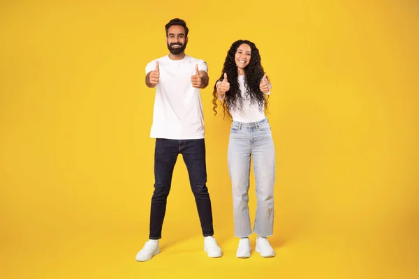 ハッピーアラブミレニアル世代男性とカメラで親指のアップと笑顔を示すカジュアルな女性は 黄色の背景の上に立っている両手でジェスチャーのように示し スタジオショット — ストック写真