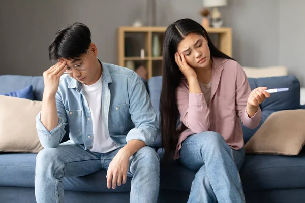 Upset asian couple with negative pregnancy test sitting on sofa at home, disappointed with bad result and reproductive health issue. Medical disorders and problems in childbearing, infertility