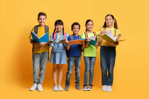 かわいい多民族学校の子供たちは元気な少年少女たちと教科書を持っているバックパックを持っている子供たち 宿題をしている子供たち 黄色い背景で隔離された完全な長さ キッズ教育 スクールコンセプト — ストック写真