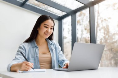 Asyalı çalışkan bayan öğrenci notlar yazıyor, makale yazıyor, web sitesi özeti yazıyor, kolej kütüphanesinde dizüstü bilgisayarla çalışıyor, masa başında araştırma yapıyor.