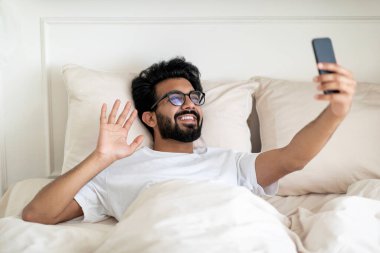 Sanal İletişim. Akıllı telefonlu mutlu Hintli adam evde yatakta dinlenirken video görüşmesi yapıyor, neşeli genç Doğu 'lu adam telefon kamerasına el sallıyor, internetteki toplantının keyfini çıkarıyor.