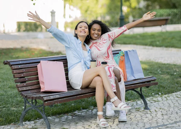 两个漂亮的年轻女人 带着购物袋坐在长椅上 快乐快乐快乐的多种族女性朋友在一起玩乐 喝着外卖咖啡 享受着季节性的销售 — 图库照片