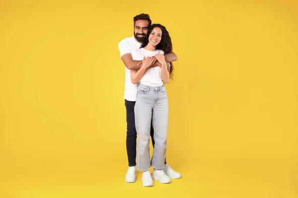 Beautiful young middle eastern couple posing embracing on yellow studio background. Handsome smiling arab man hugging his pretty wife, standing in casual outfits together. Love And Relationship
