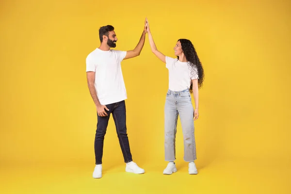 幸せな若いアラビアのカップルの完全な長さ 黄色いスタジオの背景の上に立っているそれぞれに高い5を与える 陽気な中東の配偶者はチームワークを楽しんで 共通の成功を祝います — ストック写真
