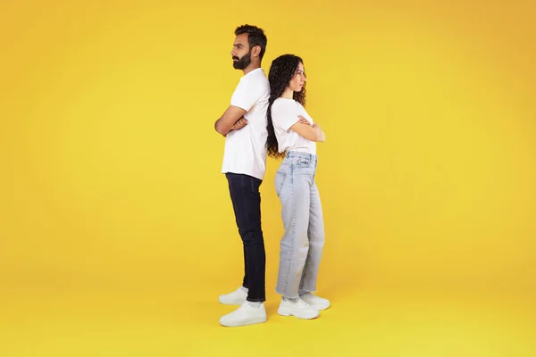 Relationship crisis, disagreement. Offended arab young spouses standing back to back over yellow background in studio, ignoring each other after quarrel, suffering marriage problems. Full length