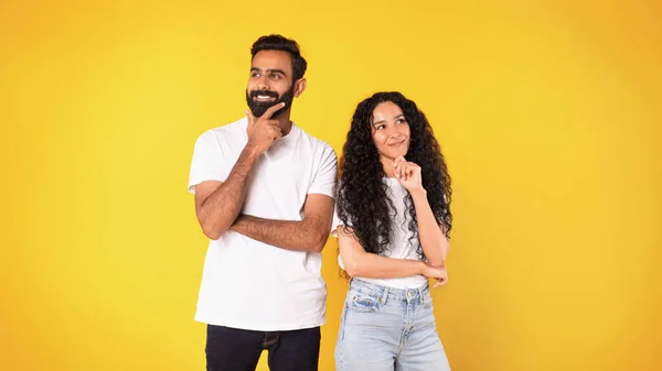 Pensive positive middle eastern couple thinking about something pleasant, touching chin posing on yellow background. Studio shot of young husband and wife dreaming looking at sides. Panorama