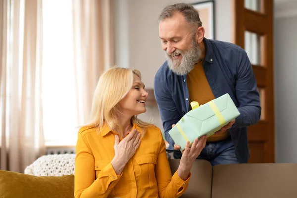 Loving senior man giving gift box to woman, making great surprise for St. Valentines Day, celebrating holiday at home interior. Husband congratulating wife on her birthday or marriage anniversary