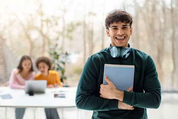 Portrait of cheerful male university student posing with notepads and smiling while his groupmates studying on background in modern audience interior