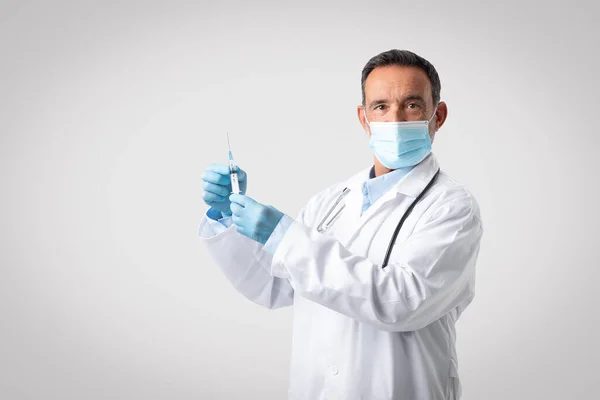 Caucasian senior man doctor in white coat, protective mask and gloves use syringe for vaccination, isolated on gray background, studio. Medicine, health care, work and treatment, immunization