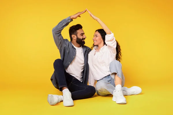 Happy Arabic Couple Sitting Joining Hands Above Head Making Symbolic Roof On Yellow Background, Smiling To Each Other Dreaming About Moving In First New Home. Mortgage for Young Family Housing