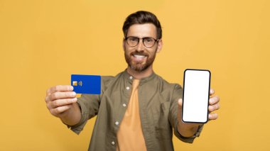 Gülümseyen orta yaşlı adam beyaz ekranlı ve kredi kartlı modern akıllı telefonu gösteriyor, sarı stüdyo geçmişi var, internet üzerinden mal ve hizmetler için ödeme yapıyor, alışveriş yapıyor veya online bankacılık yapıyor.